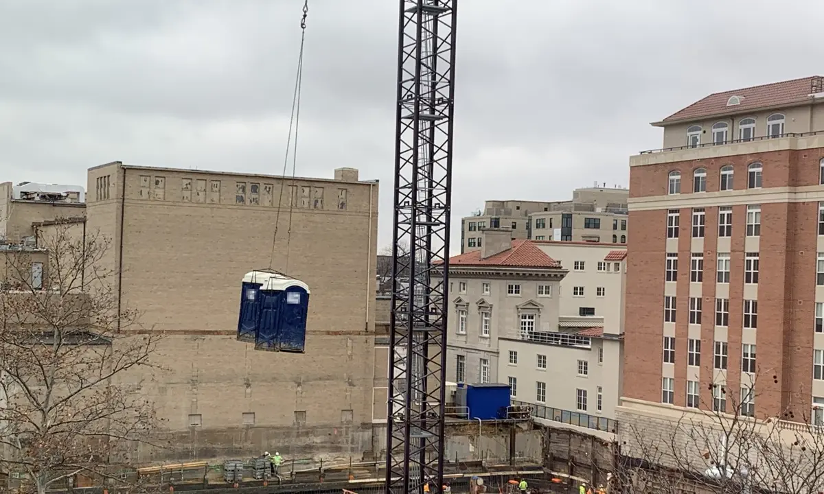Two portable toilets on a lifting frame hanging from a tower crane over a city construction site