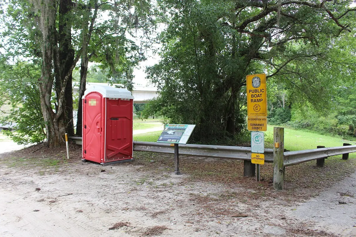 A single red standard portable toilet beside a guardrail at a public boat ramp