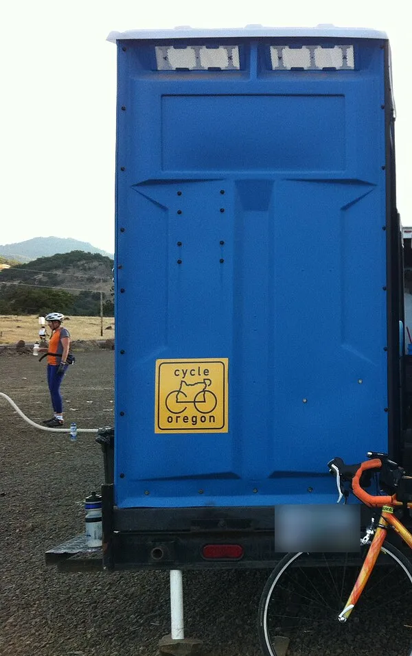 A blue portable toilet mounted on a small towable trailer at a rural rest stop